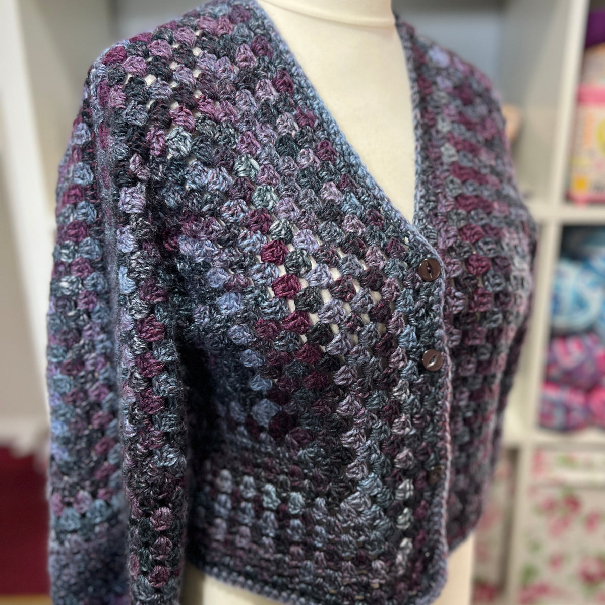 A handmade crocheted cardigan sweater displayed on a cream mannequin torso. The cardigan features a textured, variegated pattern of gray, purple, and blue yarn.