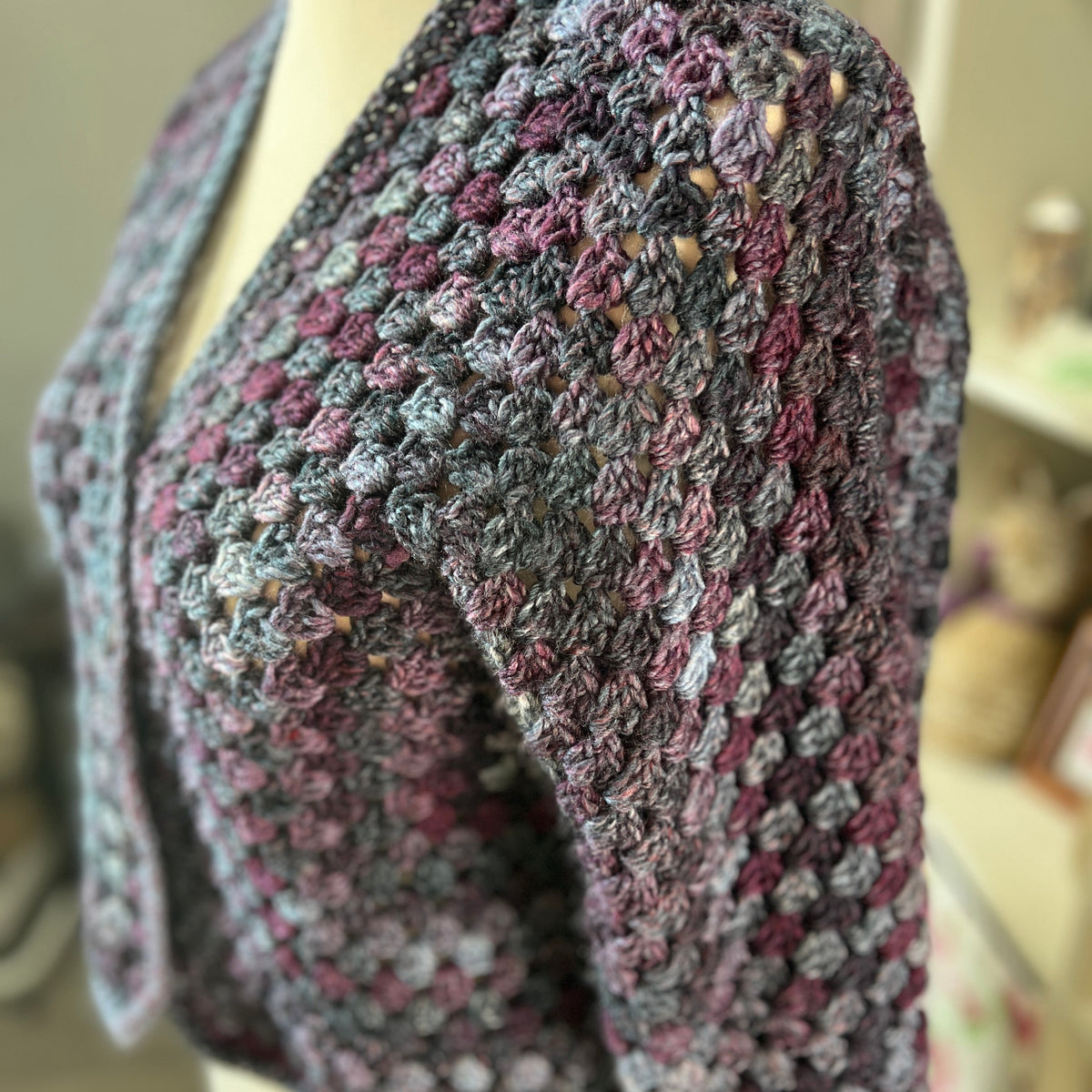 A close-up of a handmade crochet shrug or cardigan with a textured bubble pattern, displayed on a cream-colored mannequin torso. The yarn is variegated with shades of gray, purple, and dusty rose.