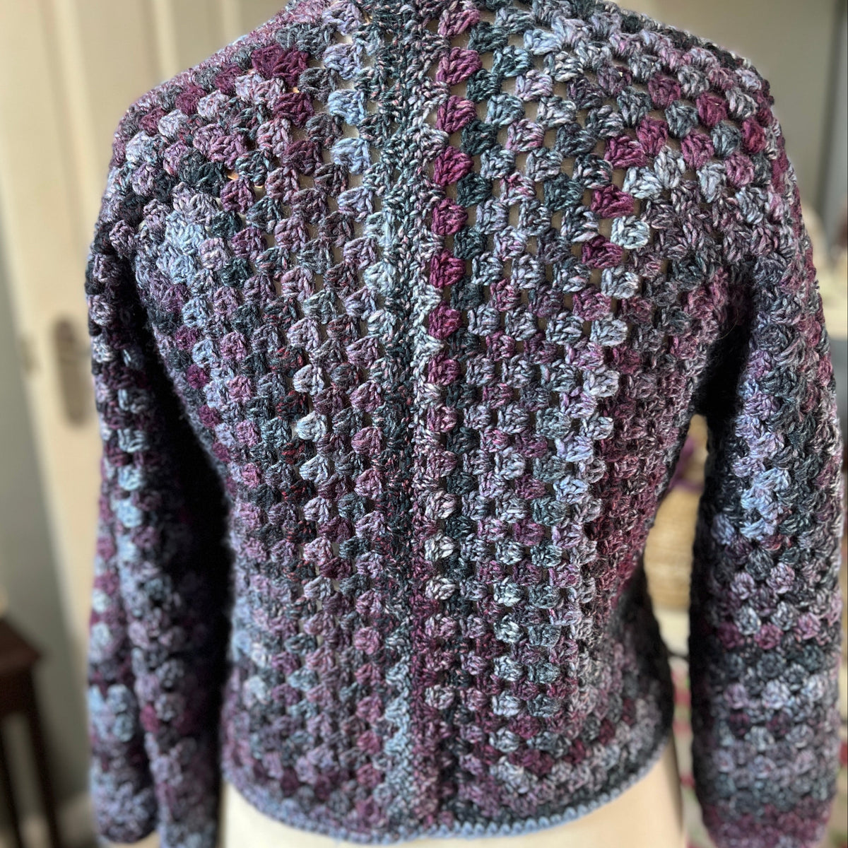 A hand-crocheted long-sleeve cardigan sweater displayed on a cream mannequin torso, showing the back view. The cardigan features a textured, variegated pattern of purple, blue, and gray yarn.