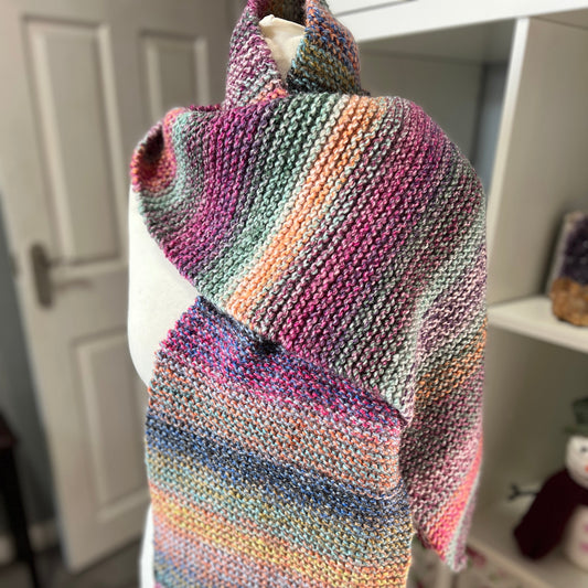 A chunky knit wrap shawl with a multicolor variegated stripe pattern, displayed on a white mannequin torso in a home setting.