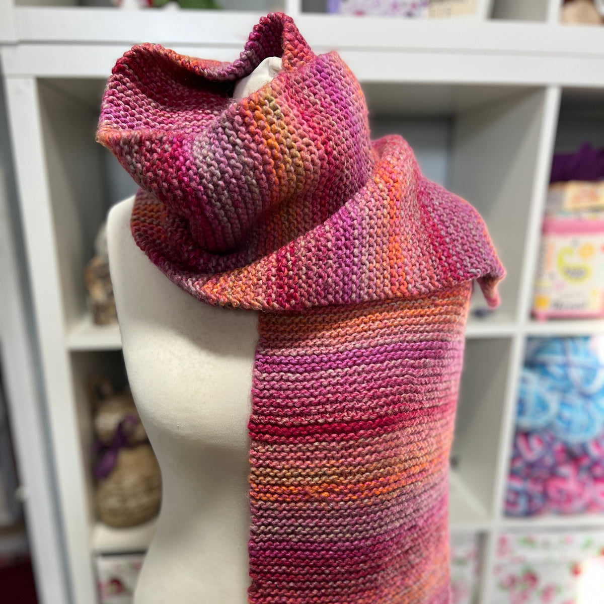 A knitted cowl scarf in a variegated pink, orange, and purple colorway, displayed on a cream-colored dress form mannequin. The background shows a white shelving unit with storage bins.