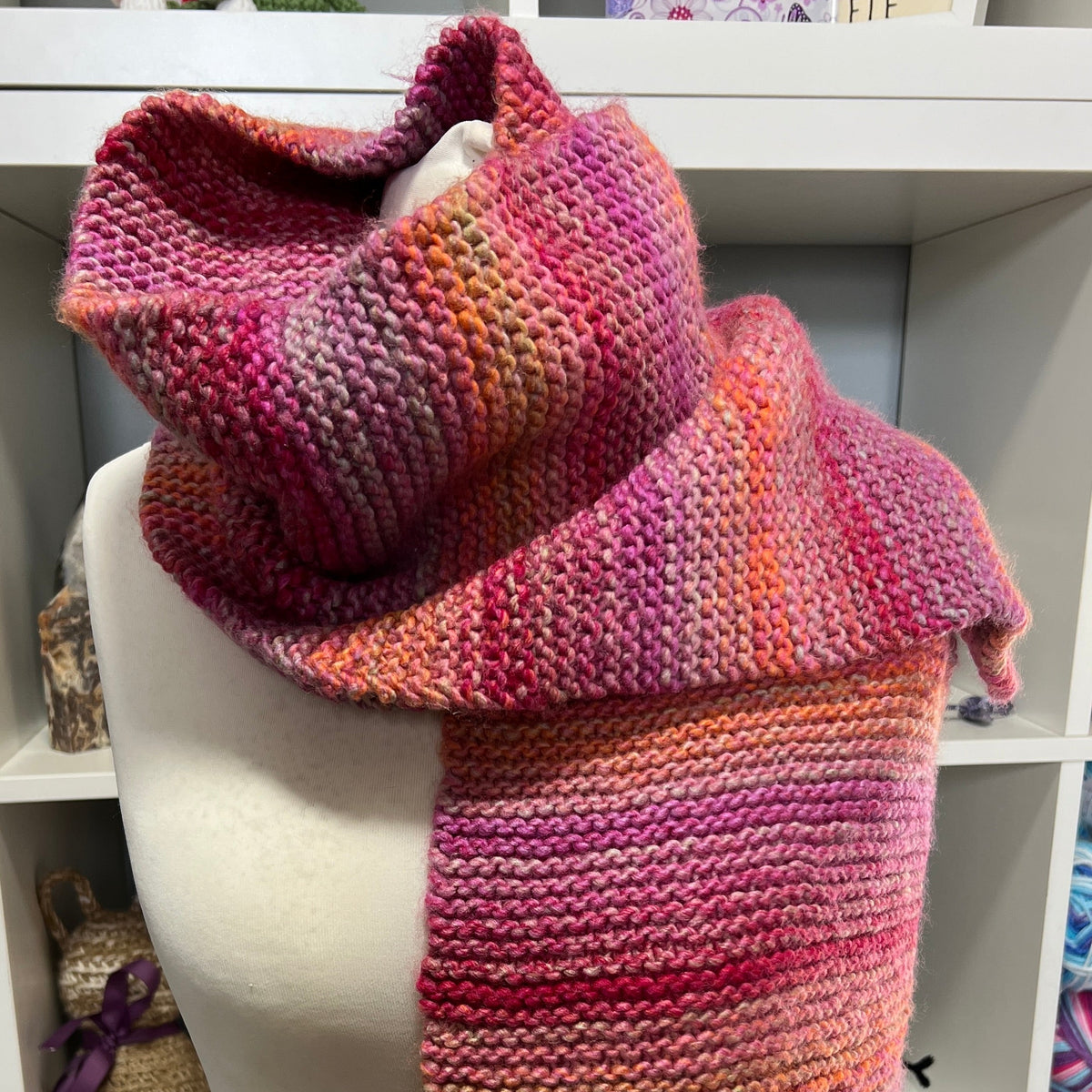 A chunky hand-knit cowl scarf in a variegated pink, orange, and purple stripe pattern, displayed on a cream-colored mannequin torso in front of a white shelving unit.