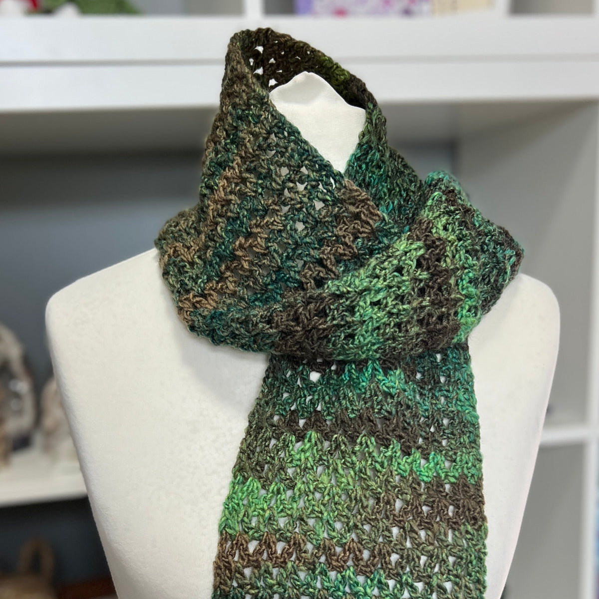 A crocheted knit scarf in a variegated green and brown colorway, displayed on a white mannequin torso in front of a white shelving unit.