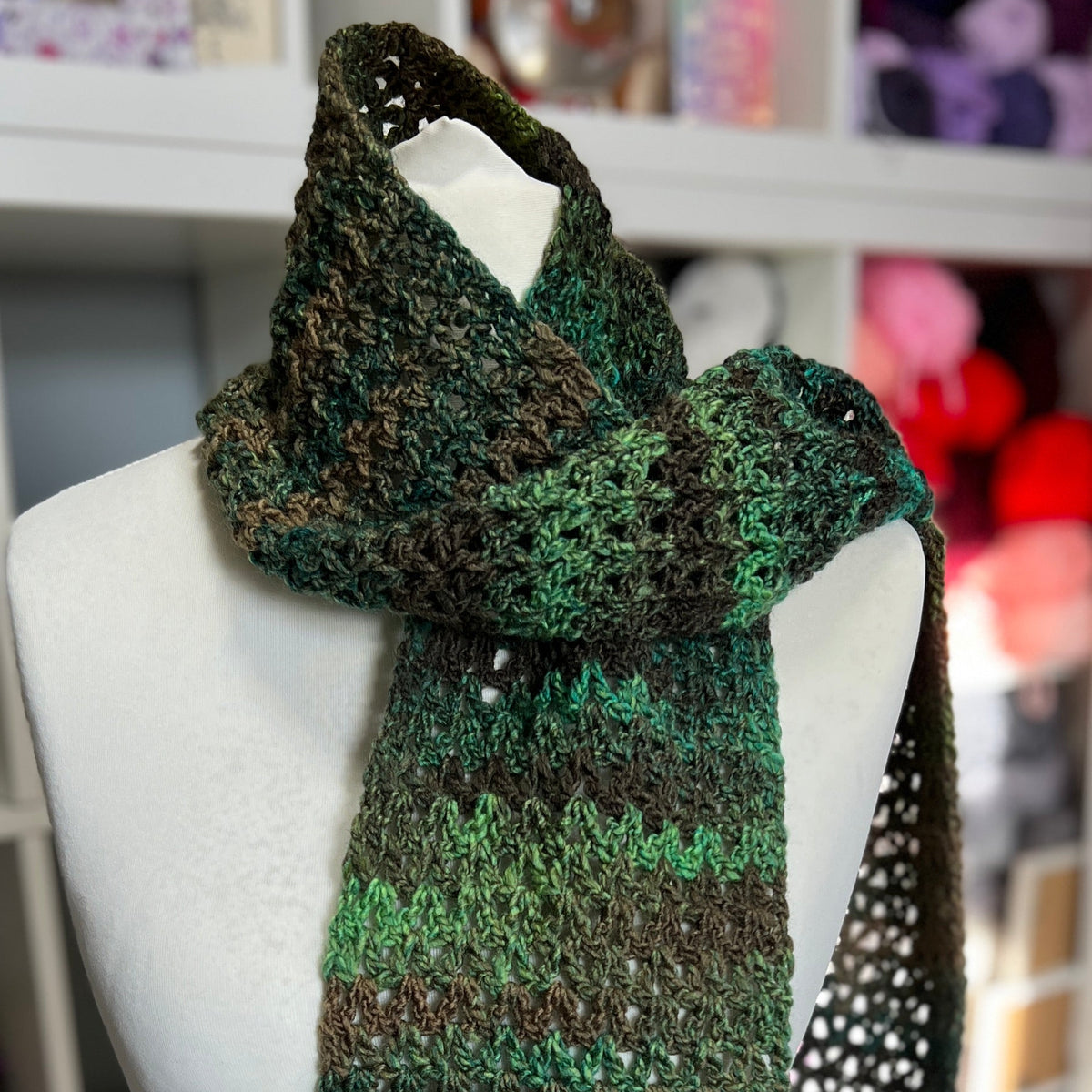 A crocheted scarf in variegated green and brown yarn, displayed on a white mannequin torso in a yarn-filled craft room.
