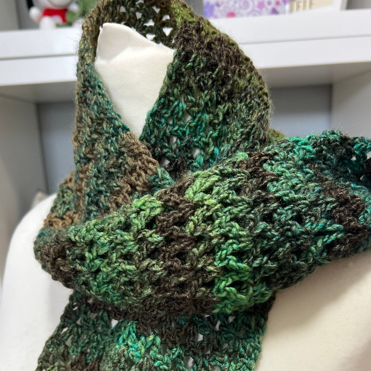 A chunky crocheted scarf in a variegated green and brown colorway, displayed on a cream-colored mannequin torso.