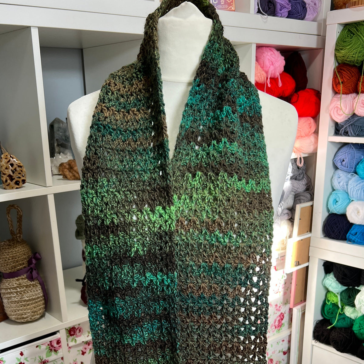 A chunky hand-knit cowl scarf with a green, brown, and black variegated pattern, displayed on a white mannequin torso in a craft room setting.